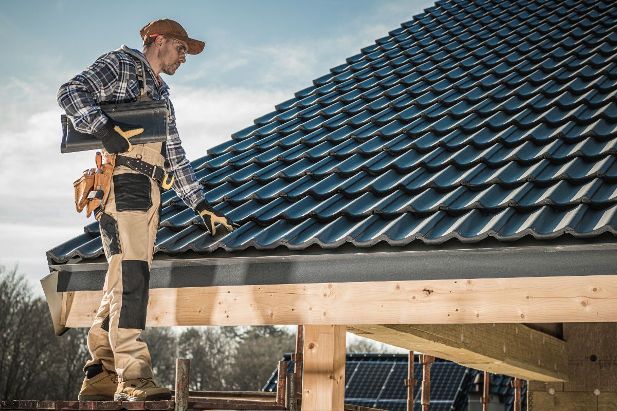 A man on a house roof holds a tool, preparing to fix a discolored roof as part of roofing services.