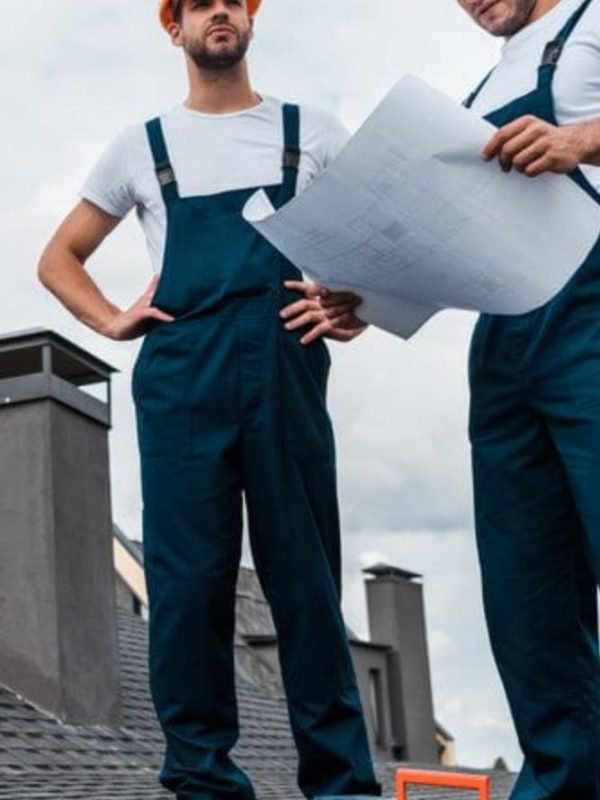 Two men in overalls standing on a roof, reviewing blueprints for a roofing project in Houston, TX.